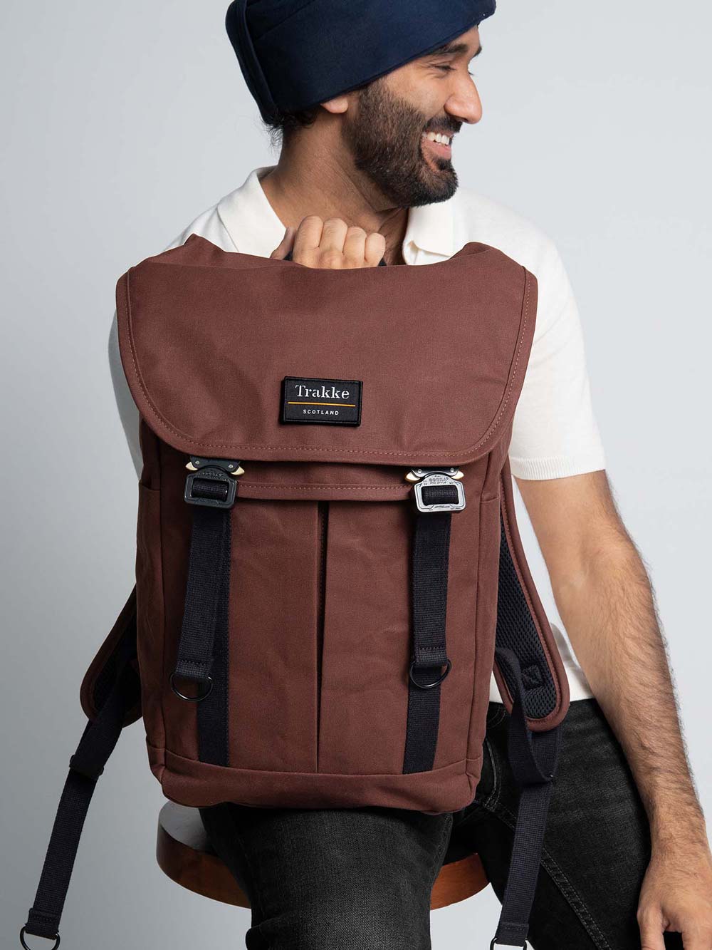 A man is sitting on a stool with the Trakke Bannoch Backpack resting on one leg. The backpack is the Port colourway and has the AustriAlpin COBRA clips.