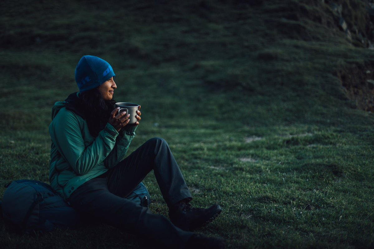 A woman drinking a hot drinking and watching the sunset, sat on her rucksack, wearing a green Jura Mountain Smock - closed
