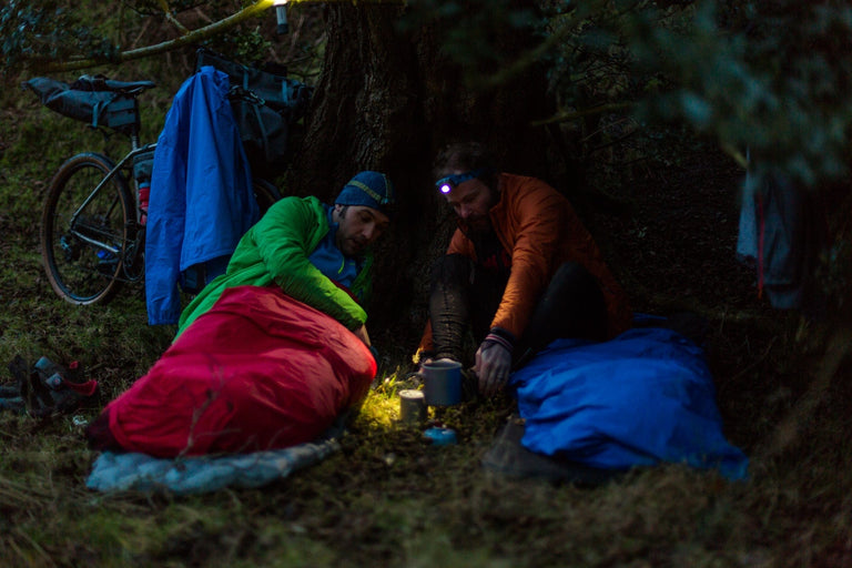 Two bikepackers cooking their tea inside their red and blue bivvy bags - action - closed