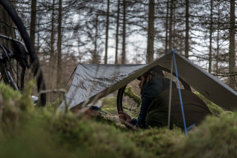 A bikepacker cooking his tea on his stove, lying down in a bivvy bag under a tarp - action - closed