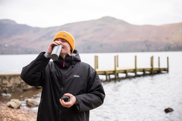 A man drinking a hot drink by Derwent water in a fleece lined swimming cloak - action - closed