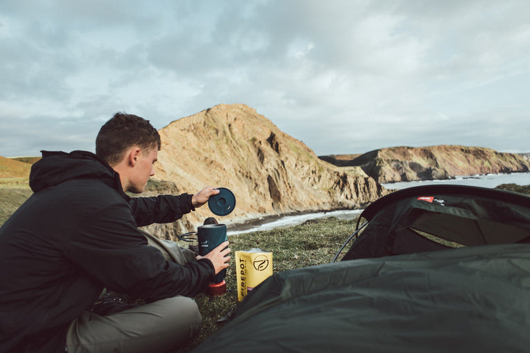 A man inspecting his stove sitting in front of his bivvy bag on the Devon coast - action - closed