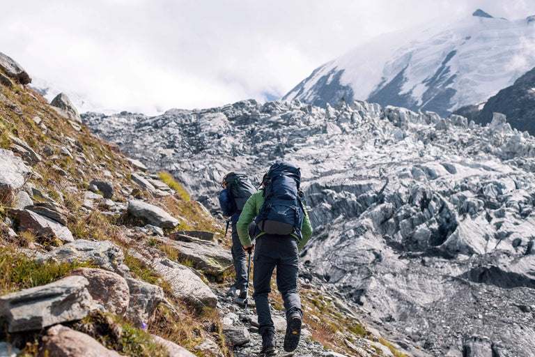 two people with backpacks and trekking poles walking up a mountain side towards a glacier - action