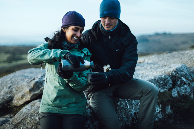 A woman and a man pouring a hot drink from a flask wearing green and black Jura mountain smocks - action