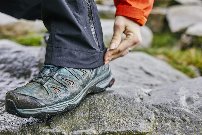 A close up of a woman in walking shoes doing up the buttons on the hem of her waterproof trousers on a rock - action - closed