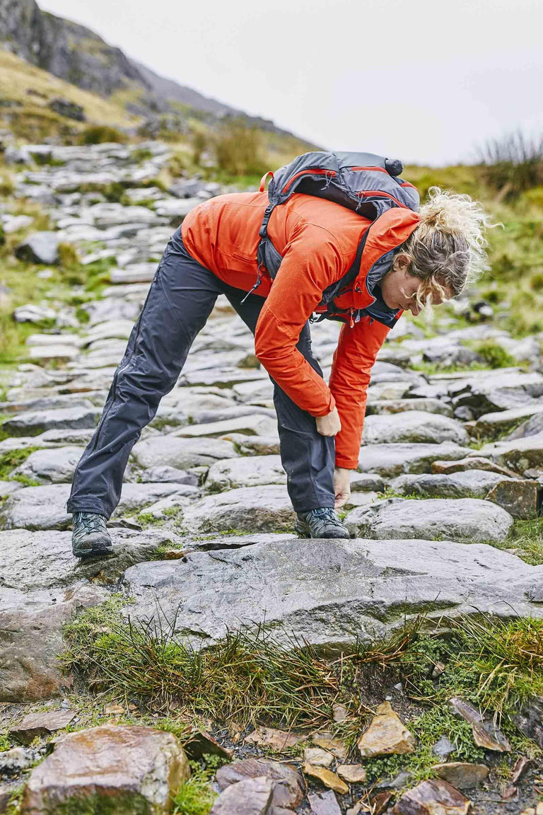 A woman in walking shoes doing up the buttons on the hem of her waterproof trousers on a rock - action - closed